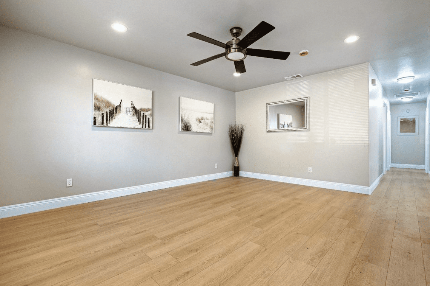 A spacious, empty room with light wood flooring, beige walls, ceiling fan, recessed lights, and wall art; a hallway is visible on the right.